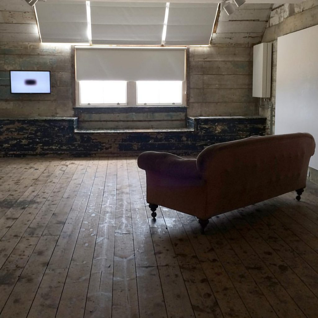 Photograph of room with wooden floor with sofa in middle and tv screen with white background image and small black square at end of room