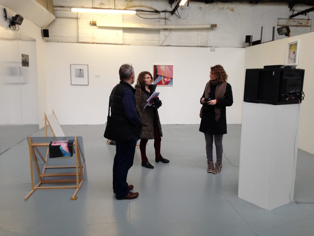 Three people in a gallery with floor based works - photographs lying on wooden A frame sculptures, colour photographs on the wall and a head height monitor.