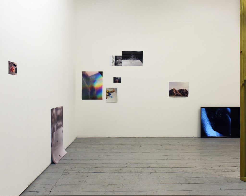 Exhibition installation shot of two walls that show various size photographs mounted directly on the wall and the corner of a video monitor.