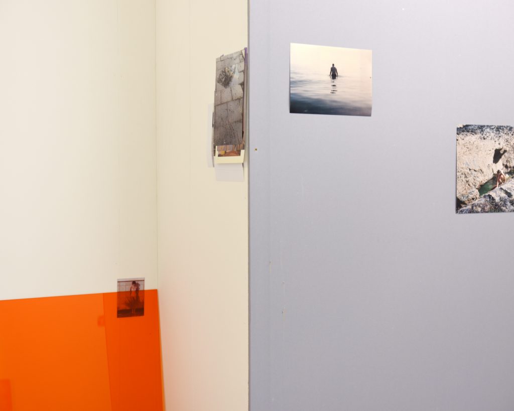 Exhibition installation shot, the corner of a room withc shows three photographs of different sizes and one small image pinned between a sheet of orange perspex and the wall.