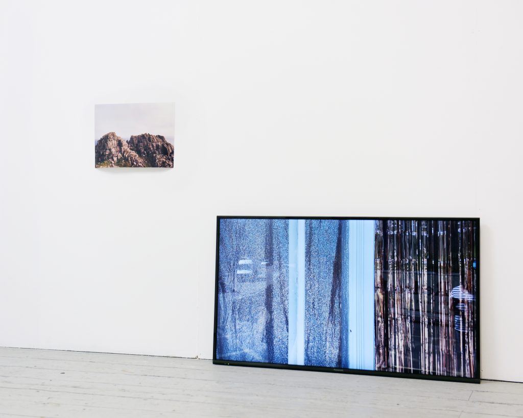 Exhibition installation photograph; photograph of rocks on left hand side on wall, video monitor leaning against wall with image of shop window with gold curtains on screen.