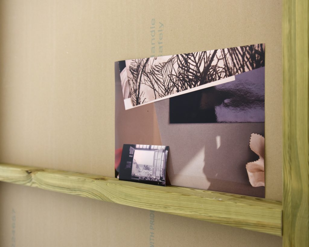 A wooden ledge with two photographs leaning against it. One of photographs layered on the floor in sunlight and one in front of it, showing the interior of a studio and the view outside.