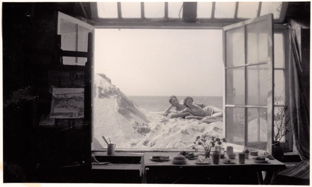 Black and white photograph taken inside an artist's studio approx. 1943, looking out through a window onto a beach where a woman and a man lie, looking at the camera.