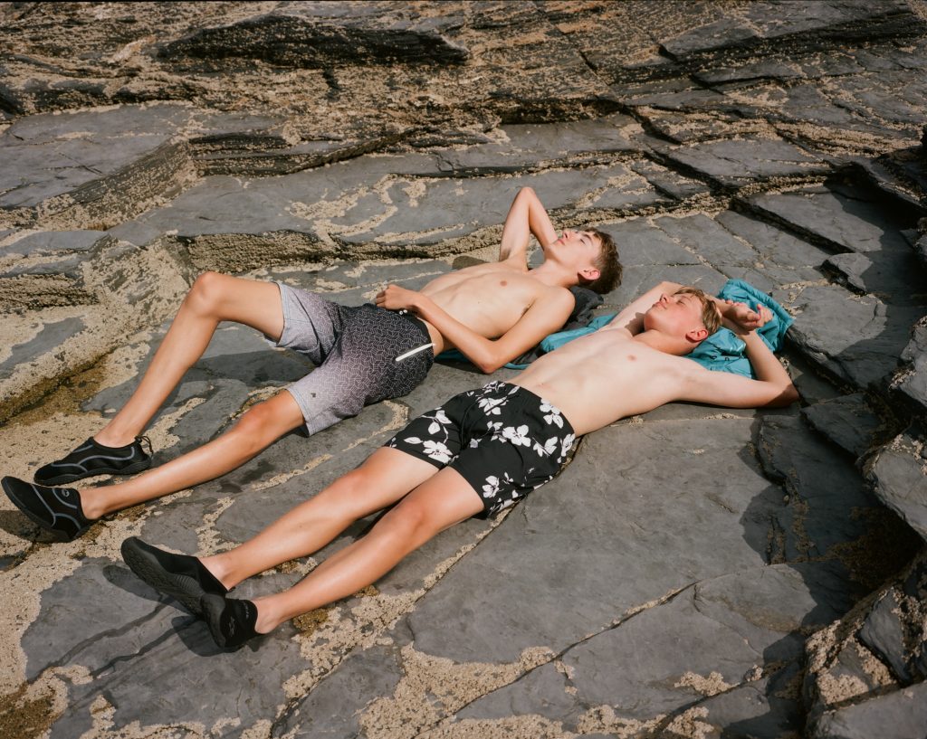 Colour photograph of two boys in swimming shorts sunbathing on rocks.