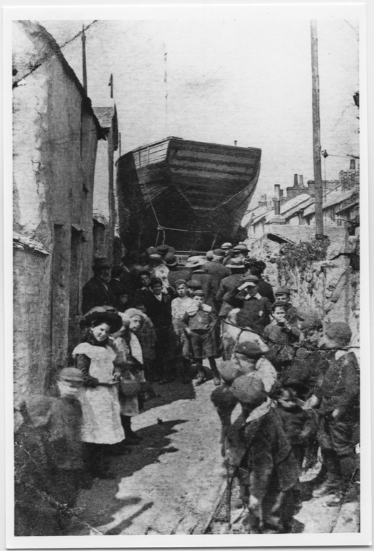 Black and White photograph, dated 1906. Penzance. Shows a narrow street full of people, facing the camera, with a large wooden boat at the back.