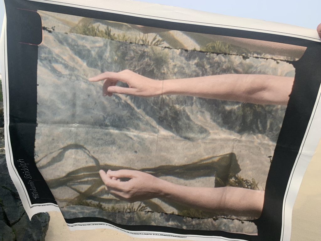 Photo of scarf on lightweight fabric, taken outdoors. Scarf features print of arms lying in water and weed.outside.