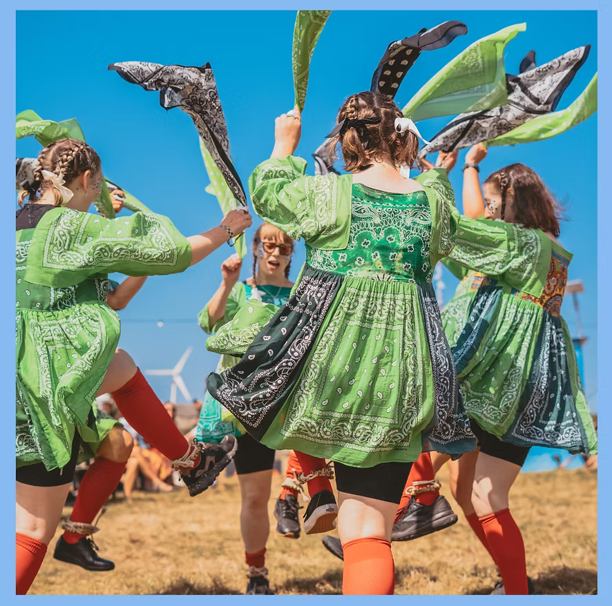 Women dancing in a circle in the sunshine with colourful green costumes and waving handkerchiefs.